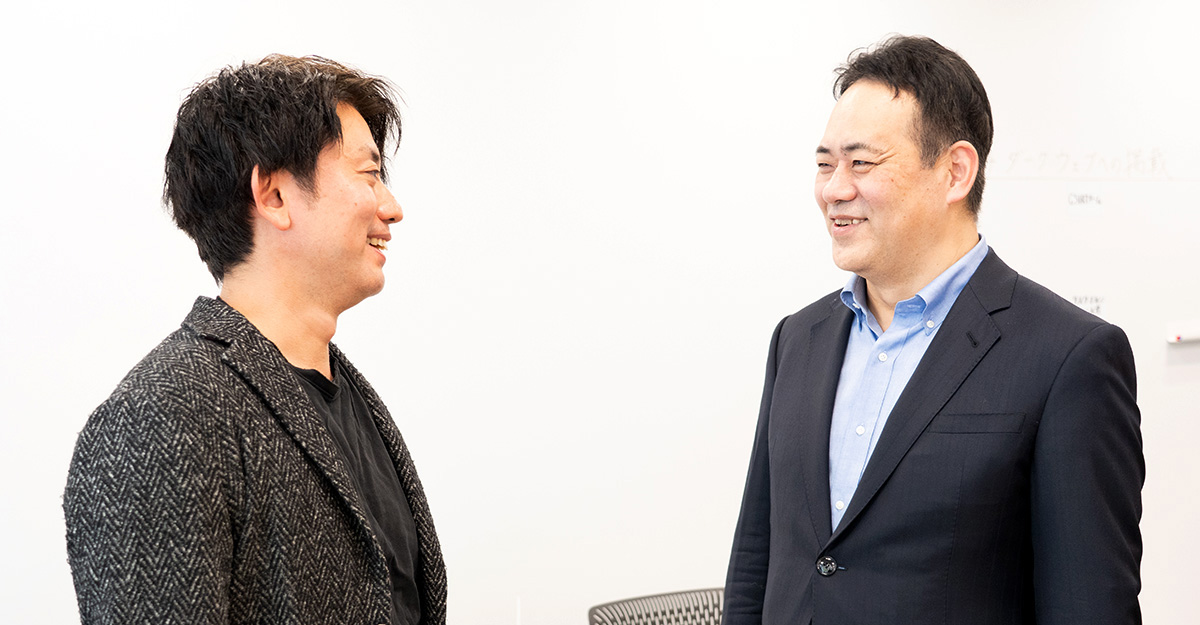 二人の男性が立ち話をし、笑顔を交わしています。背景は白い壁です。