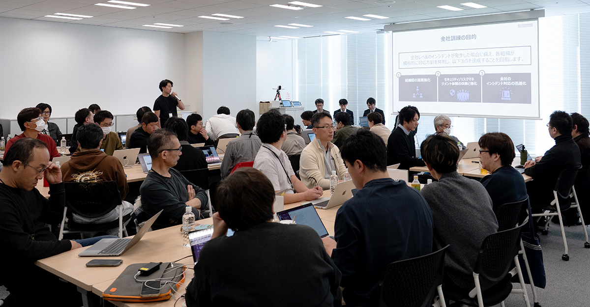 多くの人が会議室でパソコンを使いながら参加している様子。スクリーンには会議の目的が表示されています。