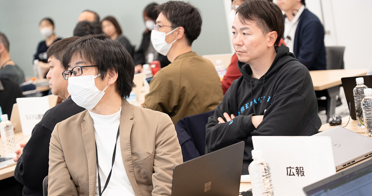 会議中の参加者がスクリーンを注視しています。机上には「広報」のプレートが見えます。