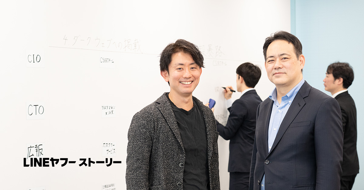 スーツを着た二人の男性が、ホワイトボードの前で笑顔で立っています。背景には他の人が書き込みをしています。