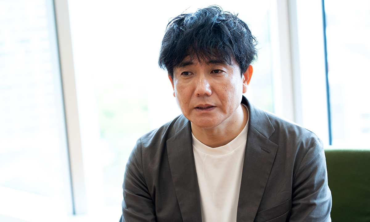 生井さんのバストショット。黒髪のミディアムヘアで、白いTシャツの上にグレーのジャケットを羽織り、やや前傾姿勢で相手の話に耳を傾けている。視線は少し下向きで、落ち着いた真剣な表情をしている。
