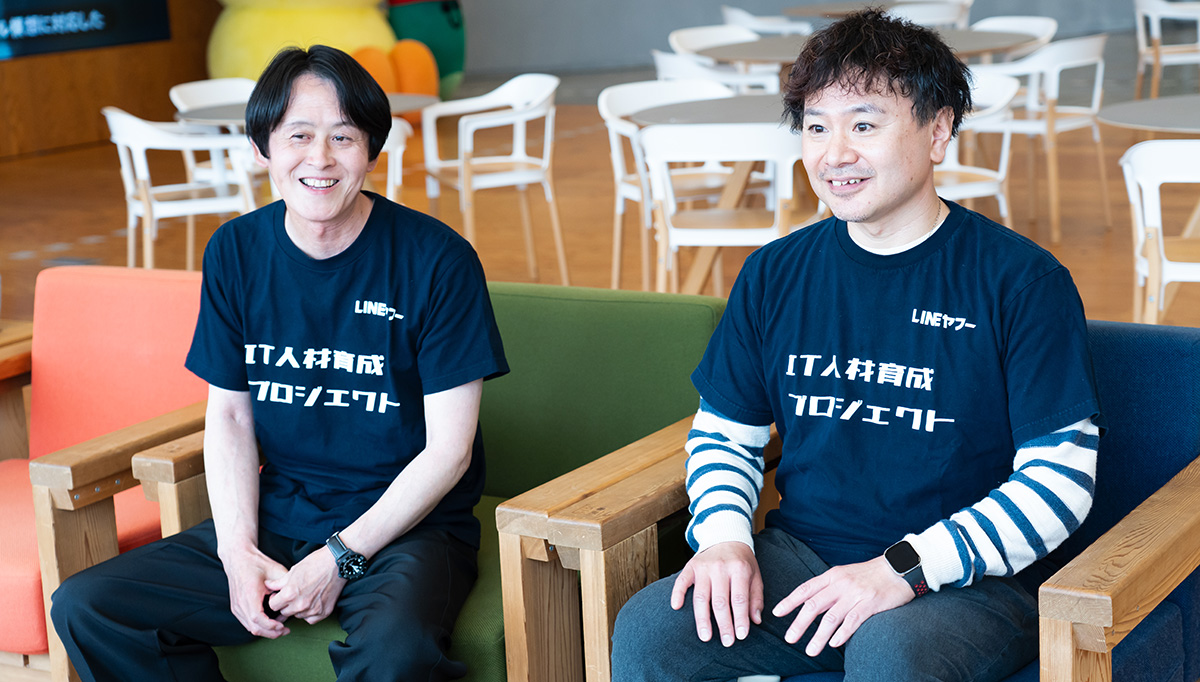 柴崎と水上のバストショット。黒い「LINEヤフー IT人材育成プロジェクト」と書かれたTシャツを着て、両手を膝の上に置きながら笑顔を見せている。