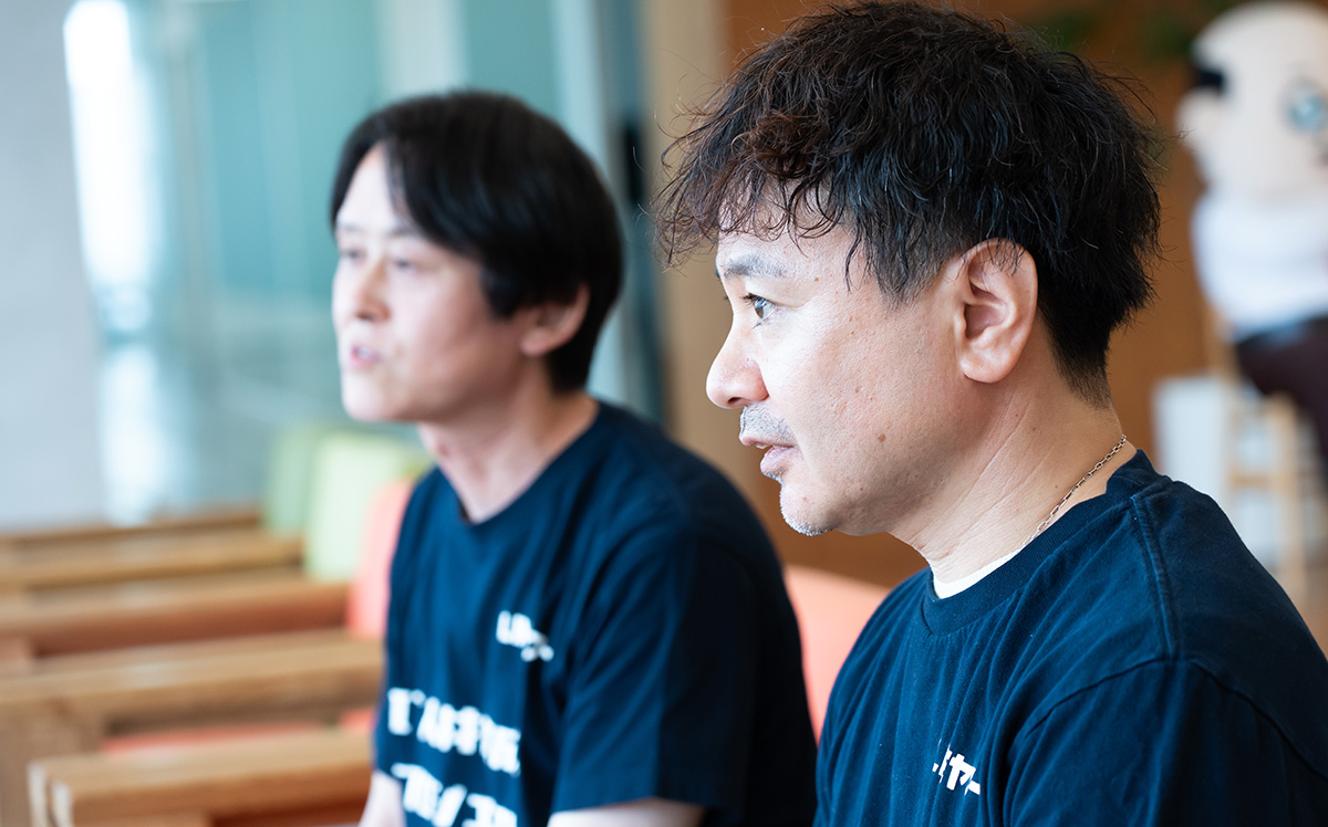 水上と柴崎のバストショット。手前に大きく写っている水上は、やや短めのパーマがかかった髪型で、黒い「LINEヤフー IT人材育成プロジェクト」と書かれたTシャツを着用し、横顔で前方を見つめながら真剣な表情で話を聞いている。