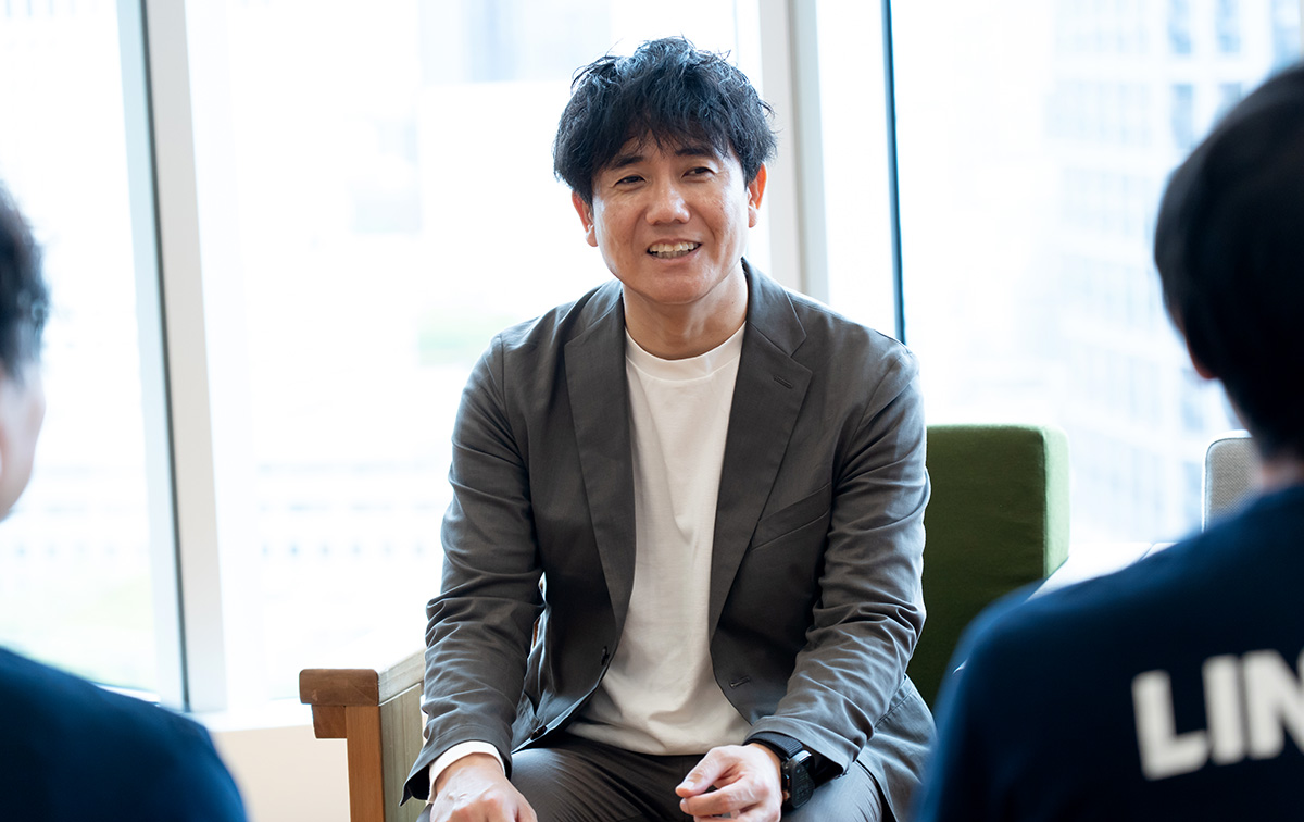 生井さんのバストショット。白いTシャツにグレーのジャケットを羽織り、木製の椅子に腰掛けている。やや前傾姿勢で両手を軽く前に出しながら、向かいに座る相手に微笑みかけている。