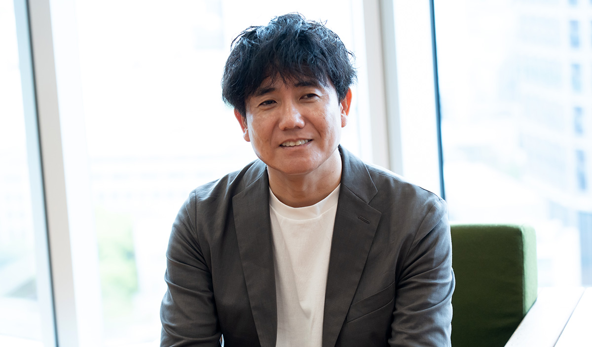 : 生井さんのバストショット。少し首を傾けながら穏やかな笑顔を見せ、前方にいる相手に視線を向けている。