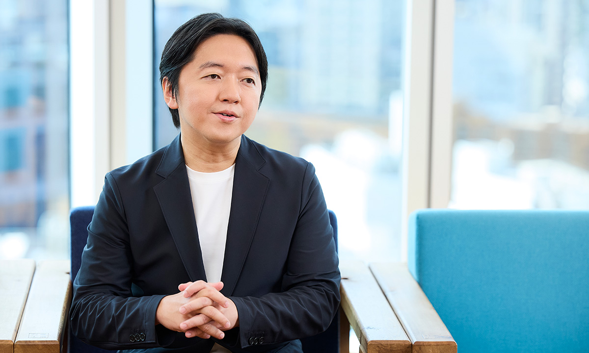 画像では、男性が座って両手を組み、話をしている様子です。背景には都会の風景が広がっています。
