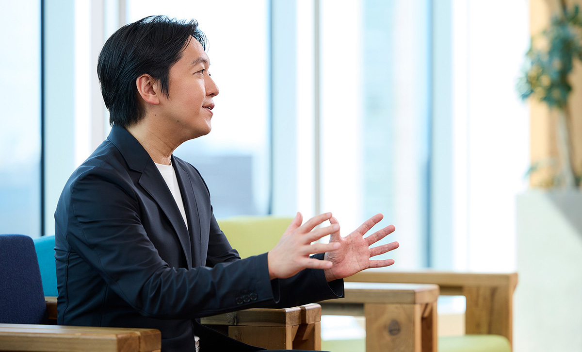 男性が横向きに座り、手を使って何かを説明している場面です。リラックスした雰囲気が伝わります。