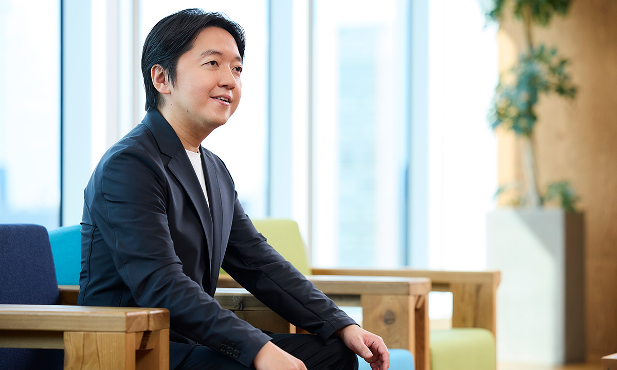 男性が椅子に座り、少し前かがみで話を聞いている様子です。背景には大きな窓と観葉植物が見え、明るい雰囲気です。