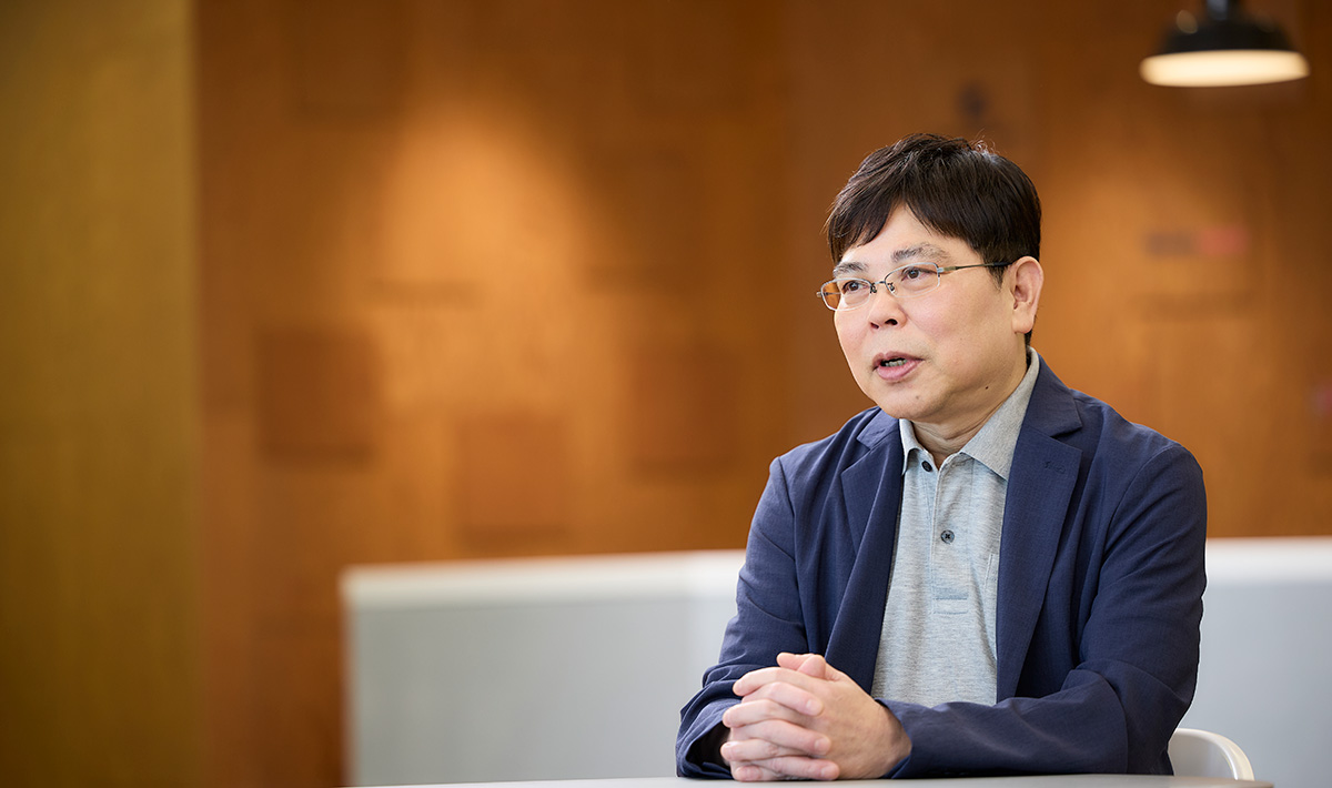 男性がテーブルに座り、真剣な表情で話しています。背景の温かみのある壁が落ち着いた印象を与えます。