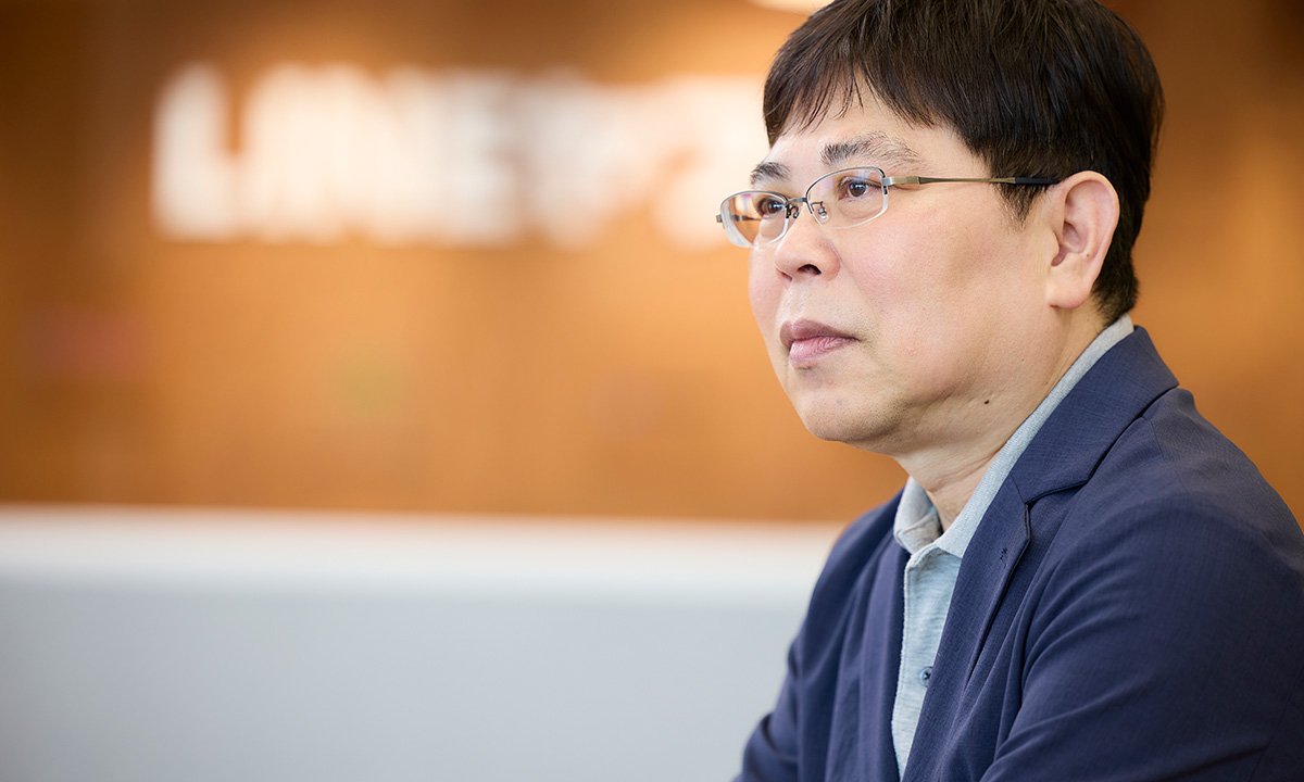 男性が真剣な表情で前方を見つめています。背景には「LINEヤフー」のロゴがぼんやりと映り、オフィスの一角が見えます。