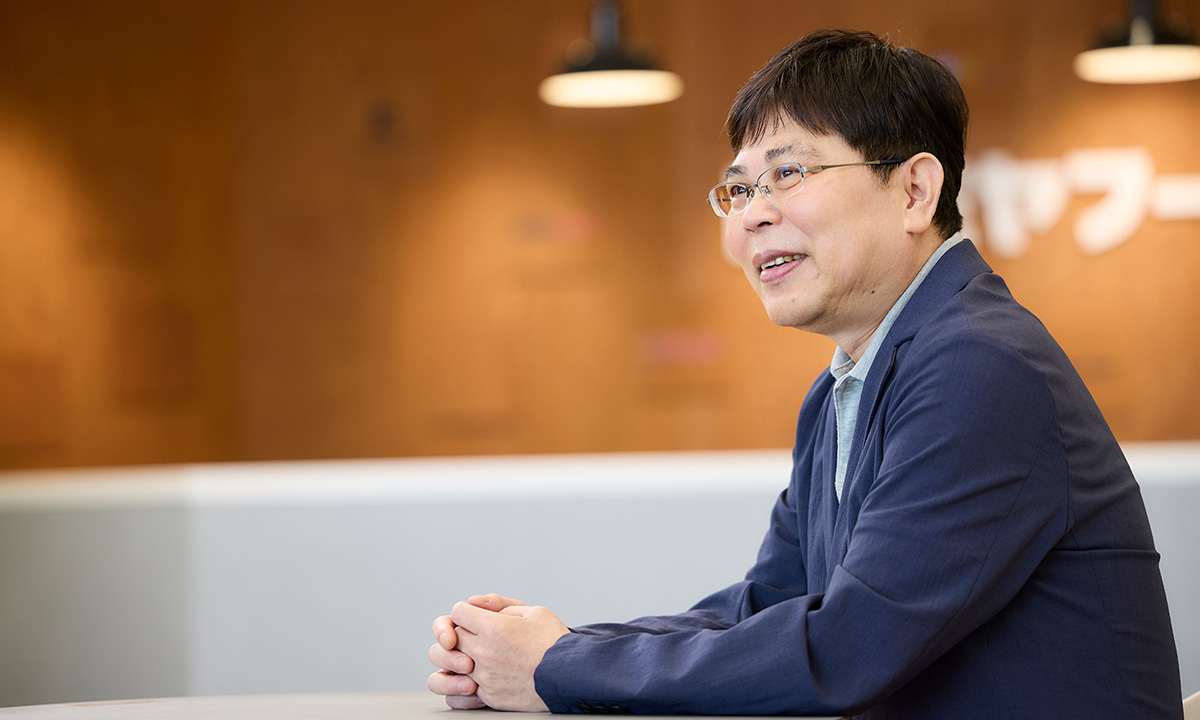 男性が横向きに座り、穏やかな笑顔で話しています。背景には温かみのある壁と照明が見え、落ち着いた雰囲気です。