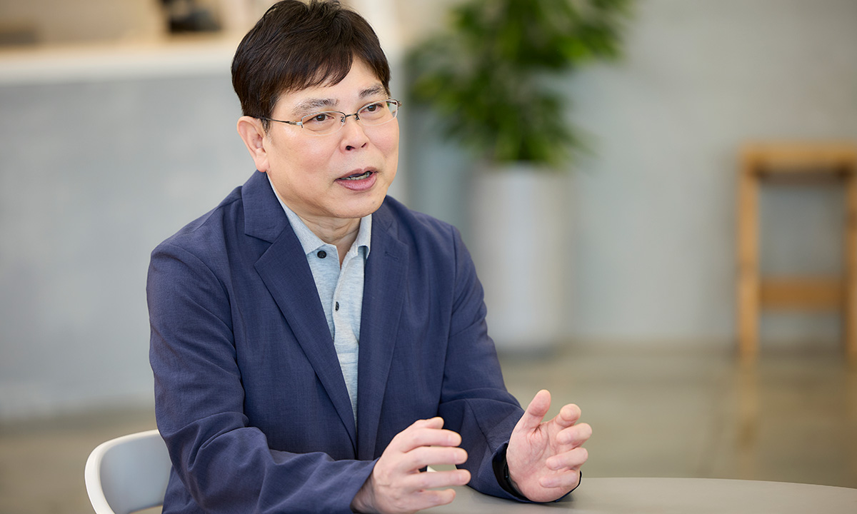 男性がテーブルに座り、手を広げて話している様子です。背景には観葉植物があり、リラックスした雰囲気が感じられます。