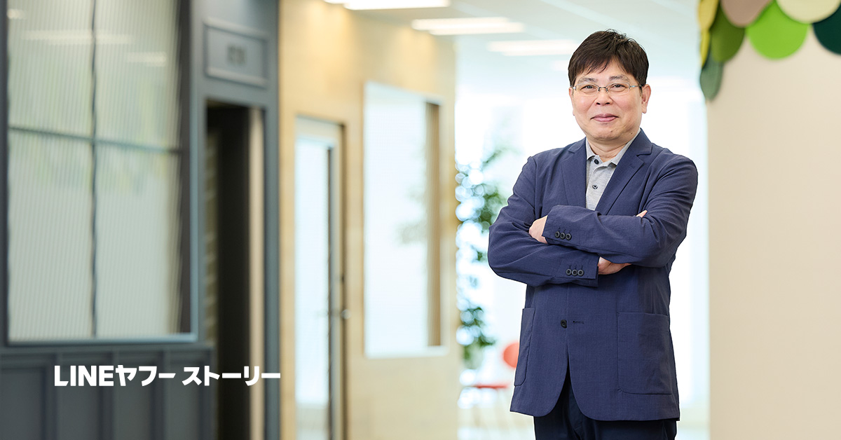 仲原がオフィスの廊下で腕を組み、微笑んでいます。背景にはガラスの扉や明るい照明が見え、洗練された雰囲気です。