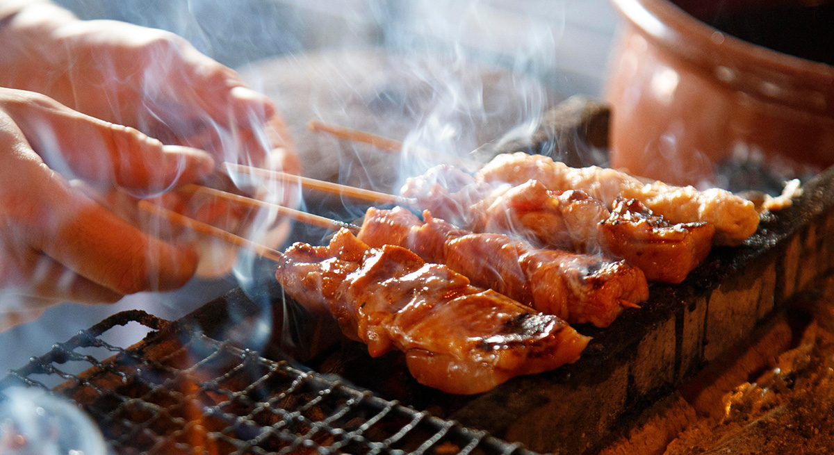 炭火の上で焼き鳥を焼いている様子。手前には照りのあるタレが絡んだ串が並び、白い煙が立ちのぼっている。