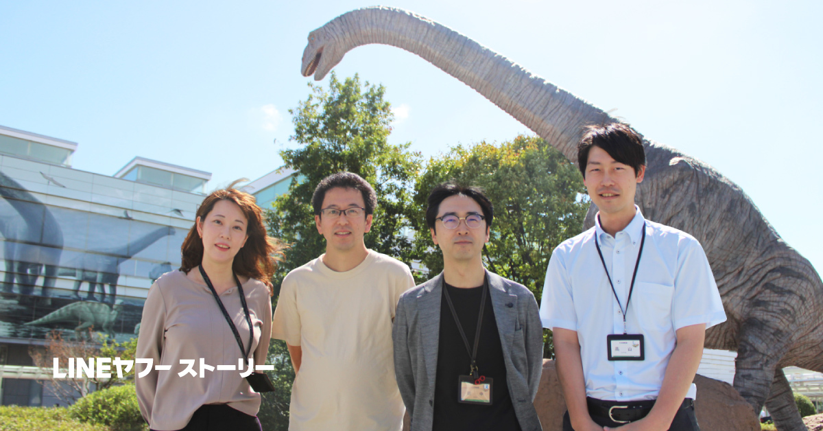 屋外で4人の男女が並んで立っている集合写真。背景には恐竜の大きな模型と「DINOSAUR KINGDOM」と書かれた建物があり、建物の壁面にも恐竜のイラストが描かれている。左下には「LINEヤフーストーリー」という白い文字が重ねられている。