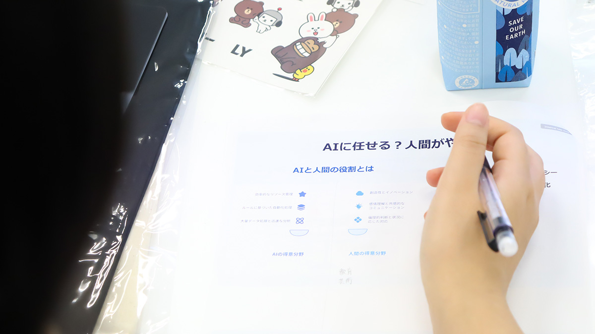 「AIに任せる？人間がやる？」と書かれた資料に、ペンを持った手が映っています。可愛いキャラクターのノートや飲み物も置かれています。