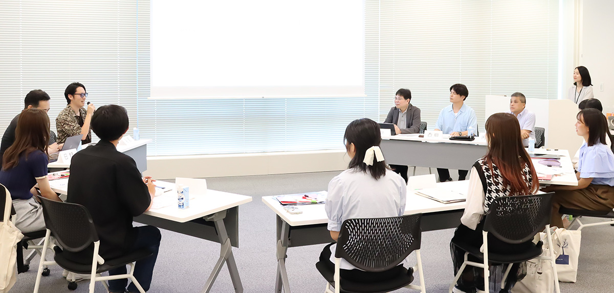 会議室で、参加者たちがテーブルを囲んでディスカッションをしています。プロジェクターに資料が映し出され、真剣な雰囲気です。