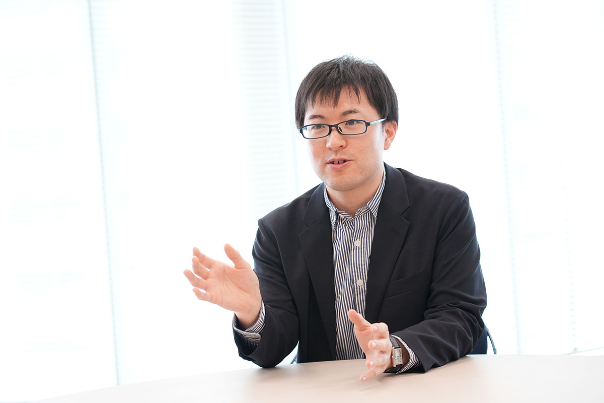 圓井がテーブルに座り、手を使って何かを説明している様子。背景には窓があります。