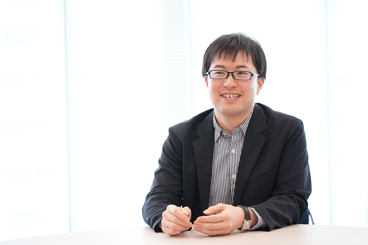 圓井がテーブルに座り、リラックスした表情で話している様子。背景は明るい窓です。