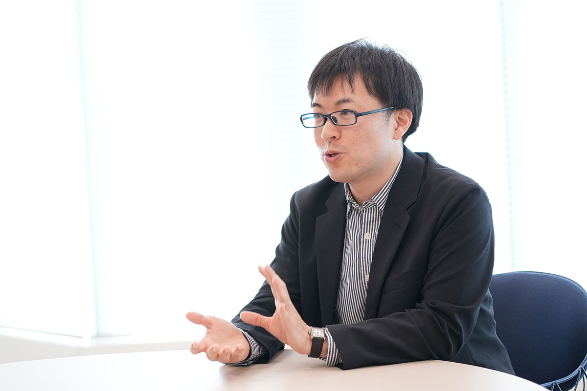 圓井がテーブルに座り、手を使って説明をする姿。背景は明るく、窓が見えます。