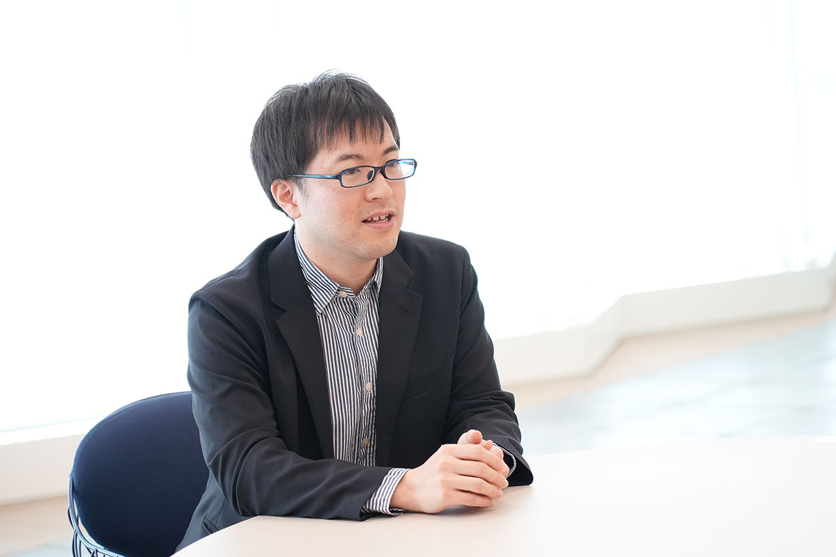 圓井がテーブルに座り、手を組んで話をしている様子。背景は明るい窓です。