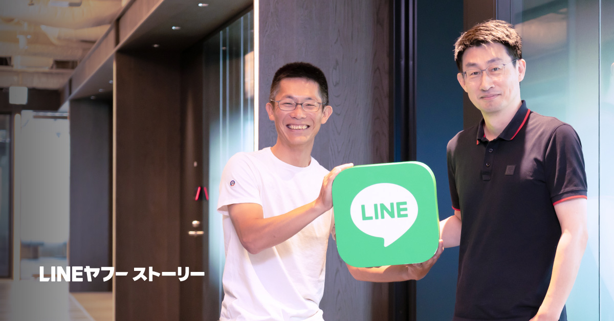 慎と朝井のバストショットです。左側の朝井は椅子に座りながら笑顔で緑色の「LINE」ロゴが描かれたクッション状のオブジェを両手で持っている。右側の慎は黒のポロシャツとグレーのパンツを着用し、短髪で眼鏡をかけ、立った姿勢で同じオブジェに手を添えながら穏やかな表情を見せている。左下には「LINEヤフーストーリー」という黒い文字が配置されている。