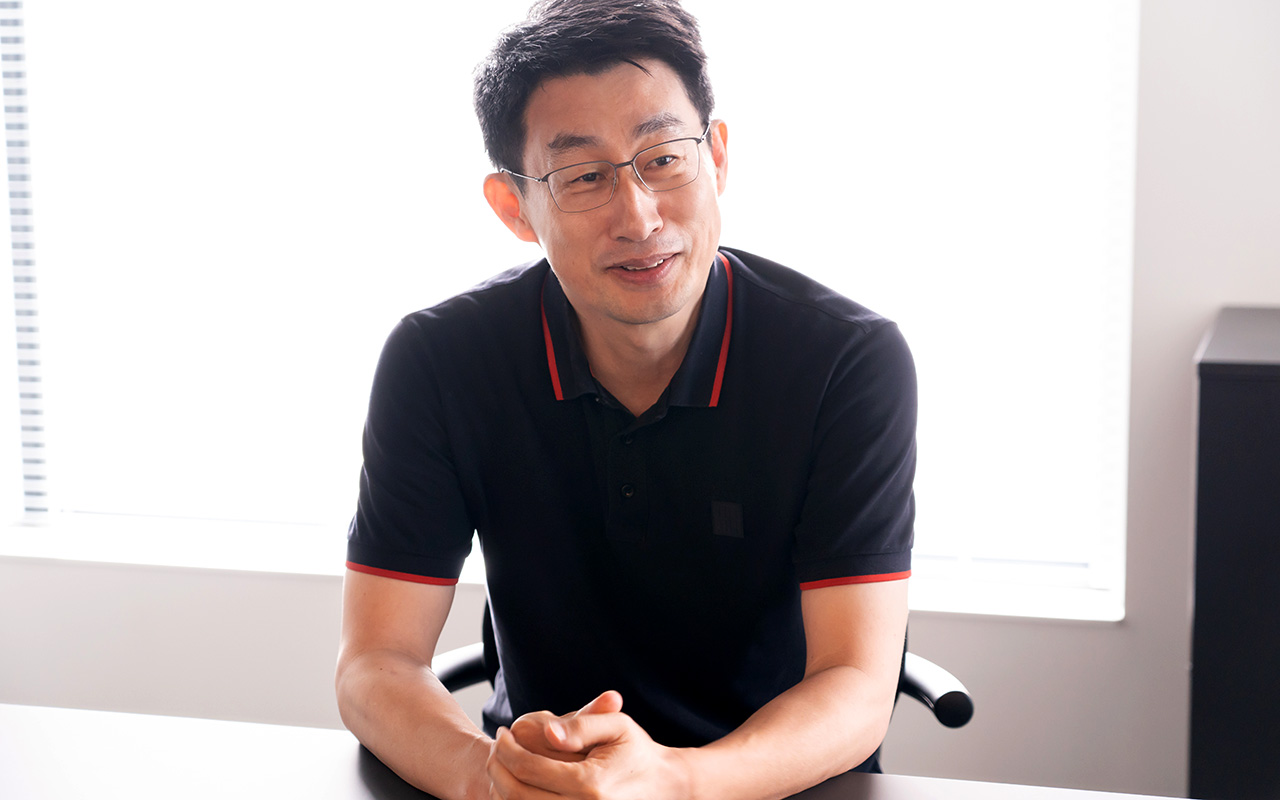 慎が、椅子に座りながら手を軽く組み、やや前傾姿勢で柔らかな表情を浮かべて会話している様子