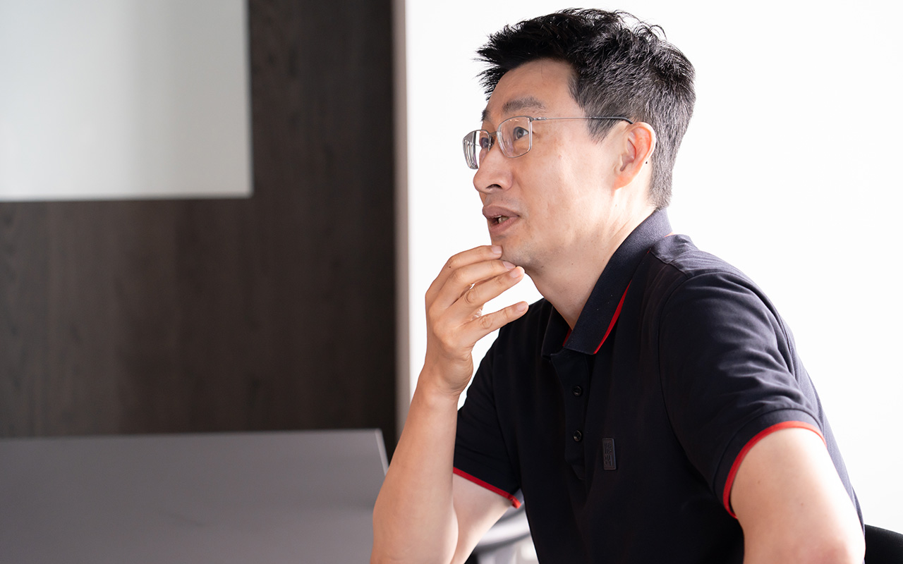 慎が片手を顎に当て、やや上方を見つめつつ考えながら話している様子