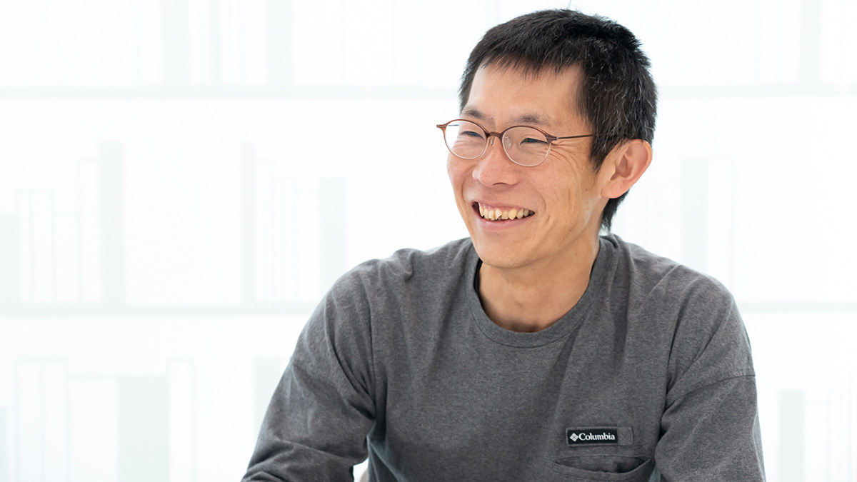 朝井が写っている写真。明るい室内で体をやや横に向け、テーブルに腕を置きながら、口を開けて自然に笑っている。背景は白を基調とした窓際で、全体にやわらかく清潔感のある雰囲気。