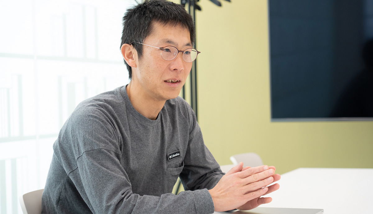 朝井が写っている写真。テーブルに向かって座り、両手をそろえて前に出しながら話している様子。視線は正面よりやや横に向けられており、真剣で落ち着いた表情で説明しているように見える。