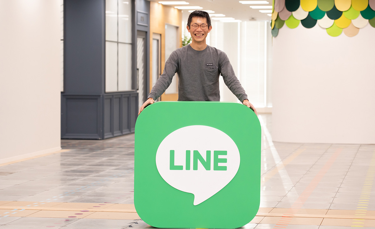 朝井が写っている写真。明るいオフィスの通路で立ち、大きな緑色のLINEロゴのパネルを両手で支えながら、口を大きく開けて笑っている。背景には白を基調とした通路やカラフルな装飾が見える。