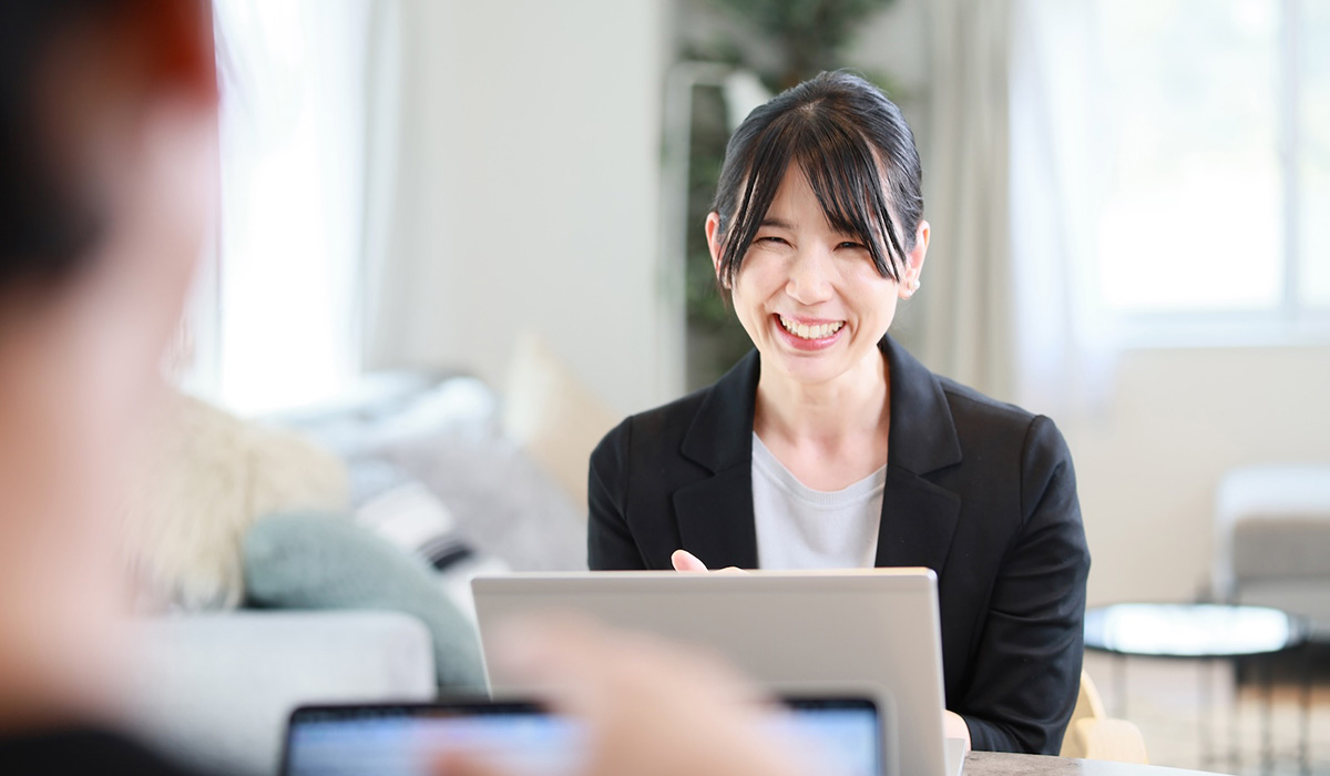 女性がパソコンの前で笑顔を見せています。手前には別の人物の手がぼんやりと映っています。
