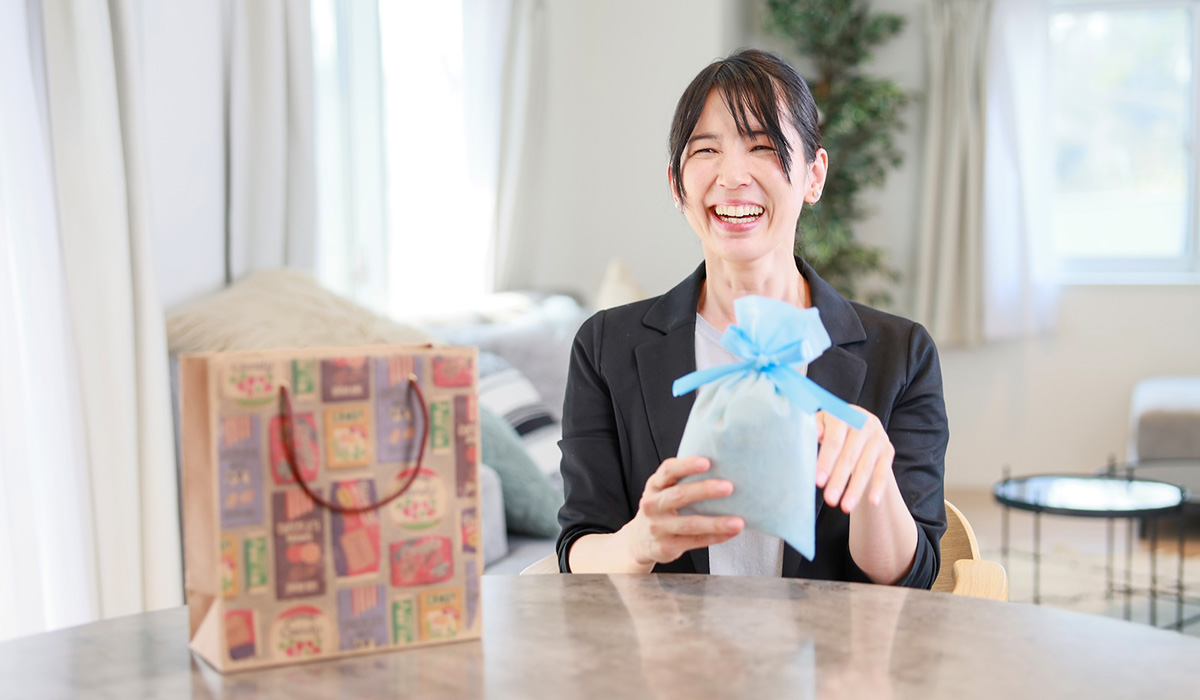 女性が青い包装のギフトを持ち、笑顔を見せています。テーブルにはギフトバッグが置かれています。