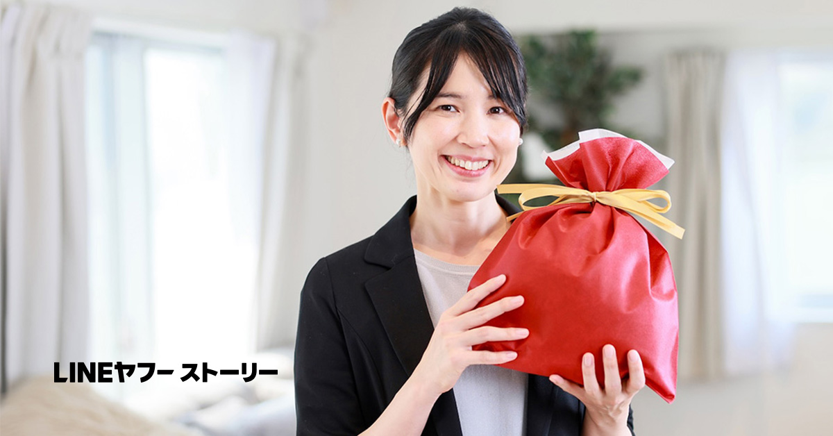 女性が赤いギフトを手に持ち、笑顔を見せています。背景には柔らかな光が差し込む部屋が映っています。