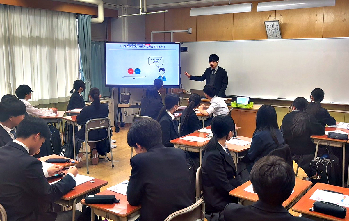 この写真は、教室で講師が生徒に説明している様子を撮影したものです。生徒たちは真剣に話を聞き、ノートを取りながら学んでいます。教育的な場面が描かれています