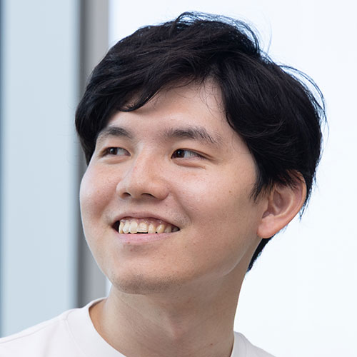 白いTシャツを着た若い男性が横を向き、口元に笑みを浮かべている写真。背景は窓と明るい外光で、柔らかく爽やかな雰囲気がある
