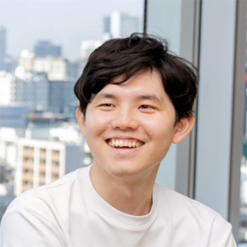 白いTシャツを着た若い男性が正面を向き、歯を見せて明るく笑っている写真。背景は白い壁で、シンプルで爽やかな雰囲気になっている