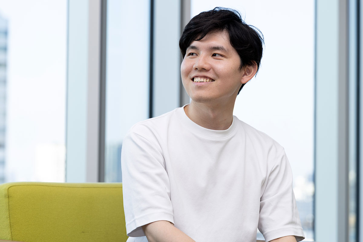 白いTシャツを着た若い男性が椅子に座り、横を向きながら柔らかい笑顔を見せている。背景は大きな窓で明るい都会の景色が映っている