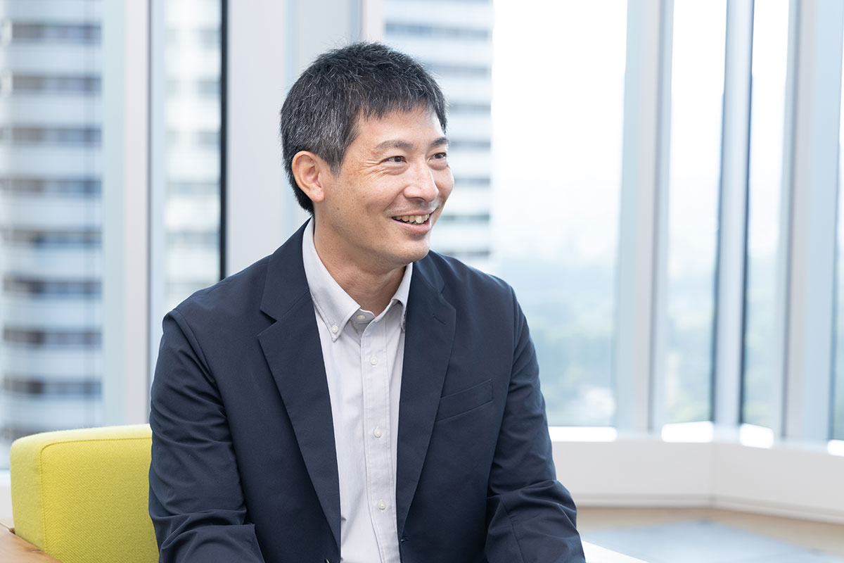ネイビージャケットを着た男性が椅子に座り、横を向きながら笑顔で会話している様子。背景には大きな窓と都会の景色が広がっている