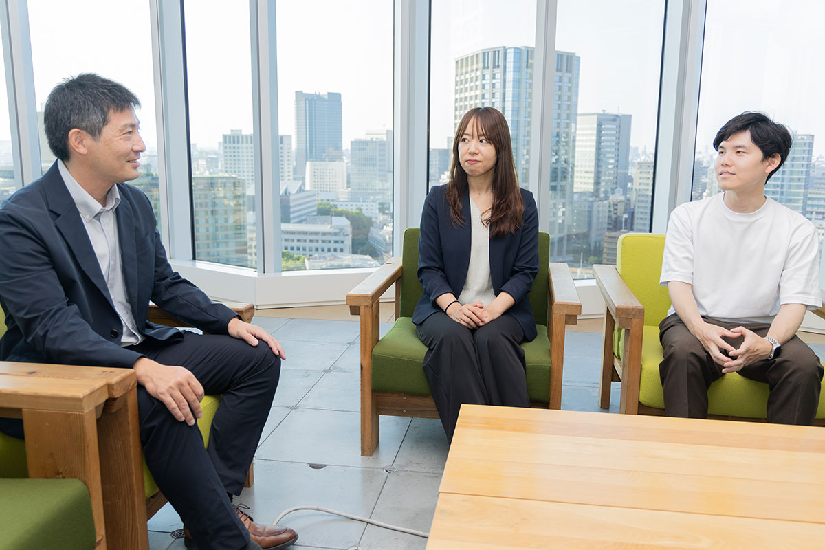 男性、女性、若い男性の3人がオフィスのソファに座り、向かい合って会話している様子。背景は大きな窓から見える都会の高層ビル群