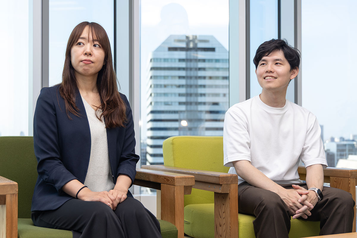 黒いジャケットの女性と白いTシャツの若い男性が並んで座っている写真。二人とも窓の外を見ており、背景には高層ビルが映っている