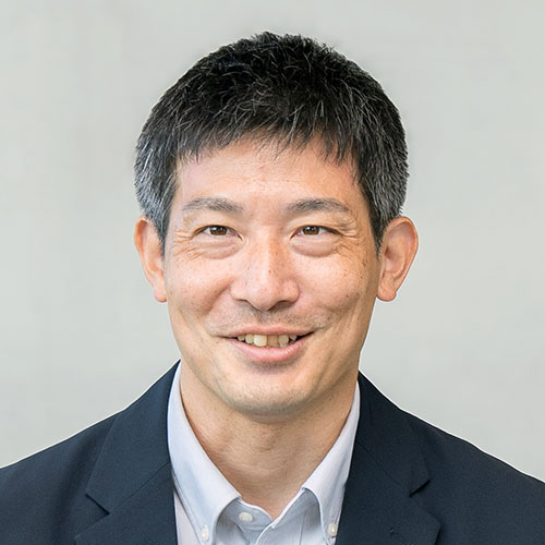 ネイビージャケットを着た男性が正面を向き、自然な笑顔を浮かべているポートレート。背景は淡いグレーで、落ち着いた印象の写真