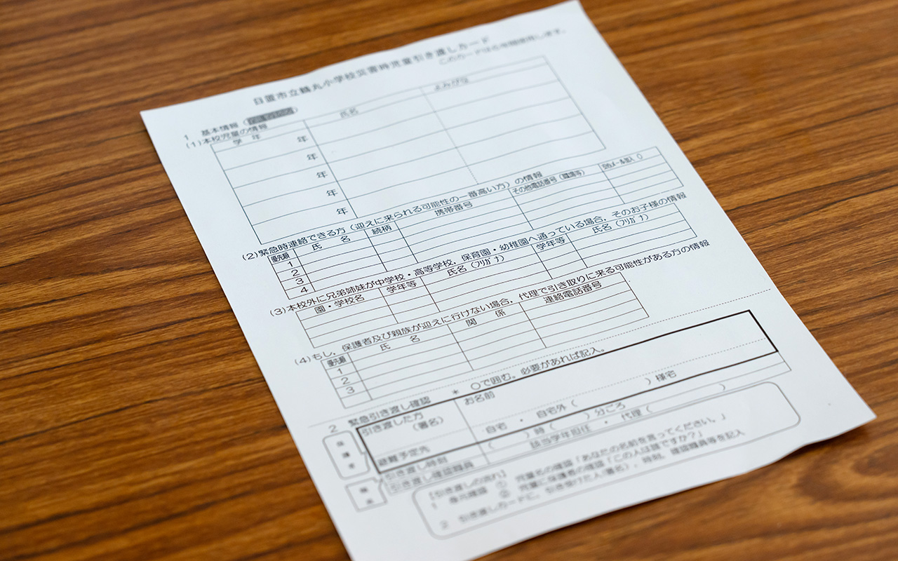 木目のテーブルの上に、記入欄が細かく区切られた白い書類が斜めに置かれている様子の写真。書類には氏名や学年、連絡先、緊急連絡先などを記入するための表形式の枠が並んでおり、日本語の見出しや説明文が印刷されている。