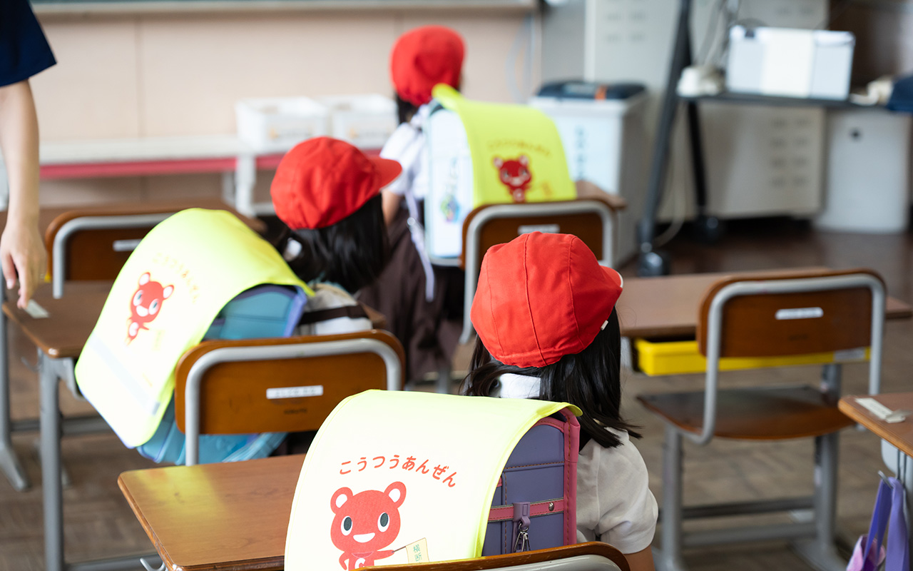 教室内で、赤い帽子をかぶった小学生たちが机に座って前を向いている後ろ姿の写真。子どもたちはランドセルを背負っており、その上から黄色いカバーがかけられている。カバーにはクマのイラストと「こうつうあんぜん」という文字が描かれている。