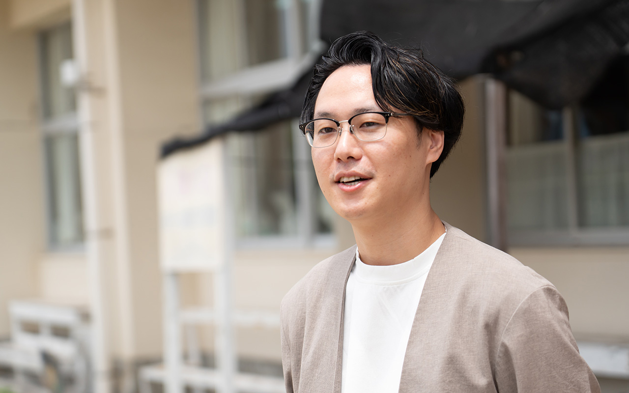 屋外の校舎前で、眼鏡をかけた高野がカメラの少し横を向きながら話している様子のバストショット。背景には校舎の壁や窓、掲示物がぼんやりと見える。