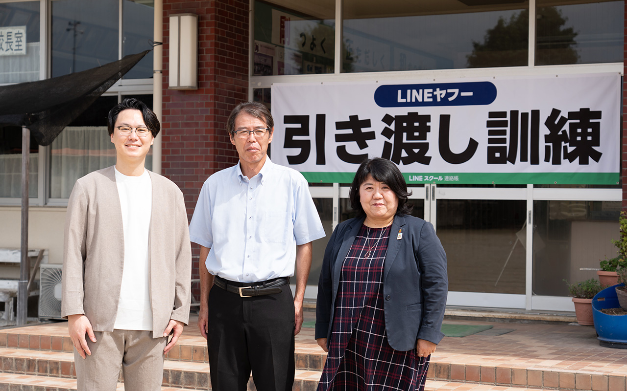 校舎の玄関前で、3人の大人が横に並んで立っている様子の写真。背後には「LINEヤフー 引き渡し訓練」と書かれた大きな横断幕が掲げられている。