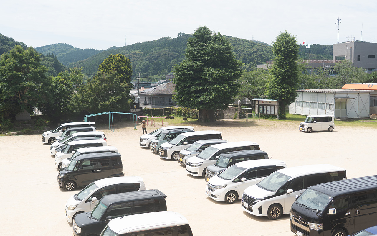 校庭に複数の車が整然と並べて駐車されている様子の写真。白や黒、シルバーのミニバンや軽自動車が列を作って配置されており、中央付近には遊具や木々がある。