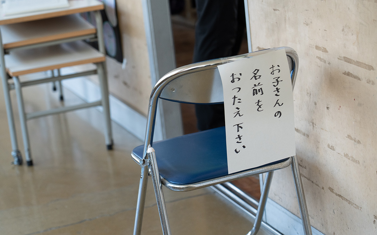 室内の床に置かれた折りたたみ椅子の背もたれに、「お子さんの名前をおつたえ下さい」と縦書きで書かれた紙が貼られている様子の写真