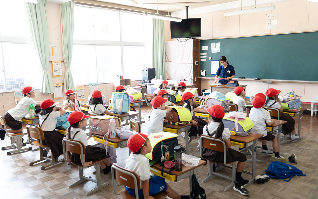 明るい教室で、赤い帽子をかぶった小学生たちが机に座り、前方に立つ女性教師の話を聞いている様子の写真。