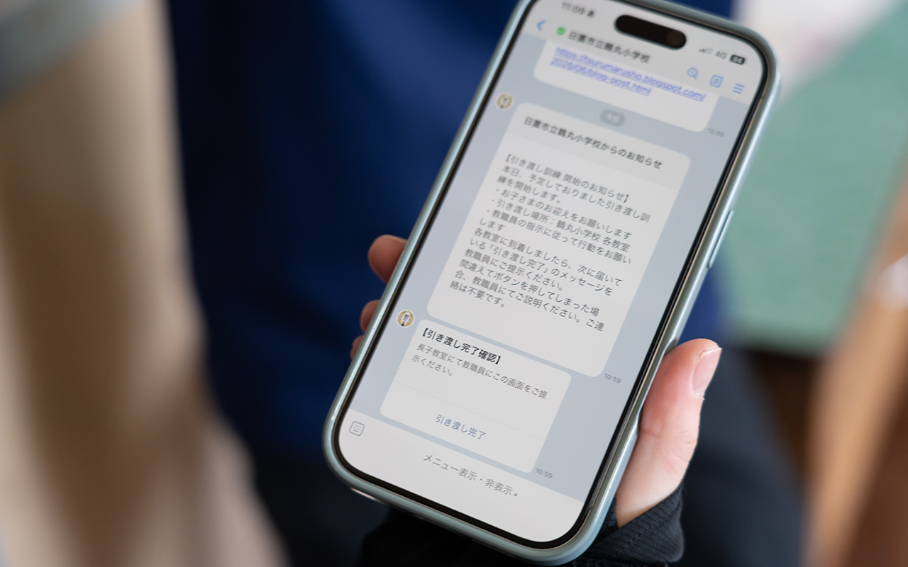 手に持たれたスマートフォンの画面に、学校からの連絡メッセージが表示されている様子の写真。画面には日本語の文章で引き渡し訓練に関する案内や確認メッセージが表示されており、チャット形式のUIになっている。